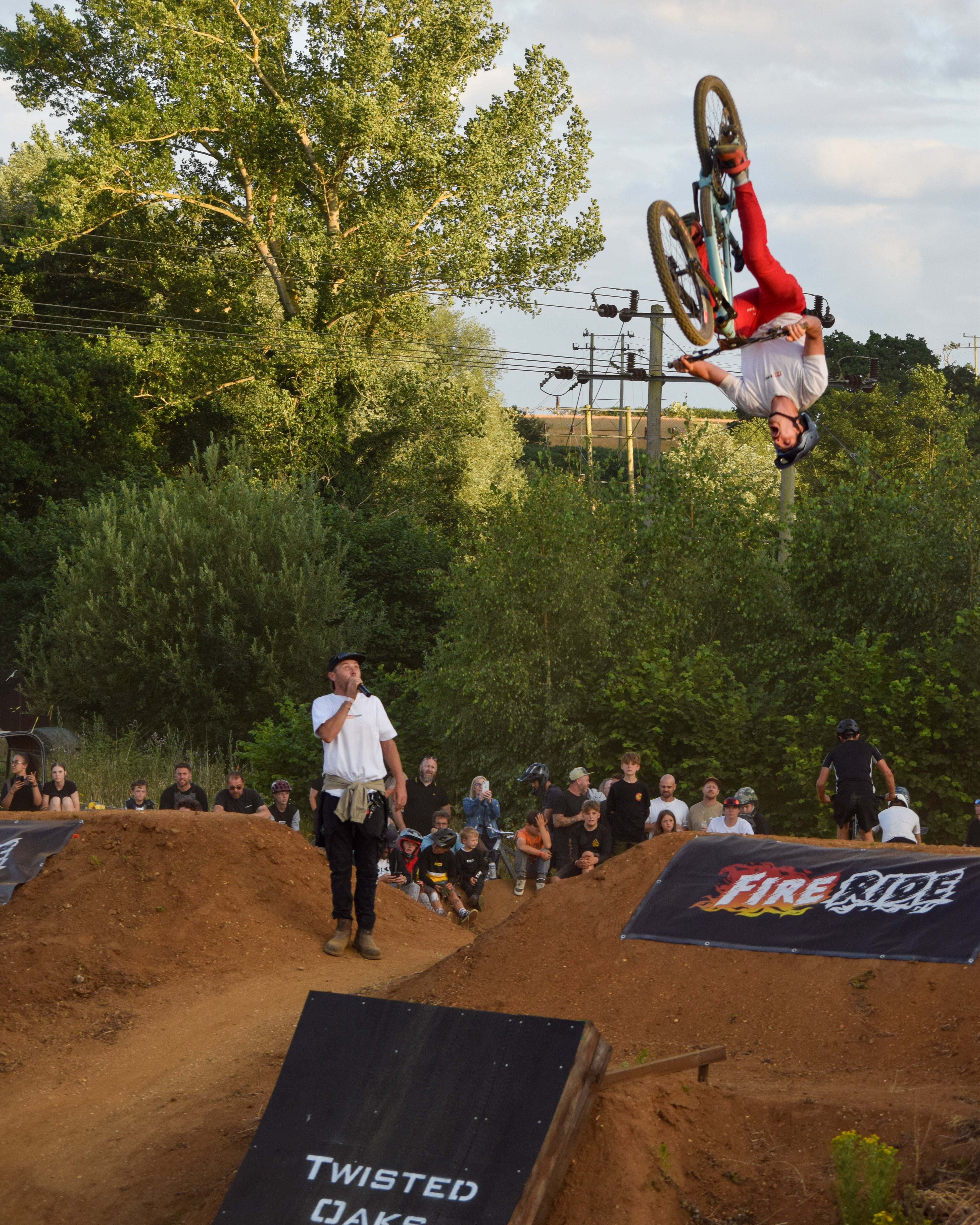 Person on a bike performing a trick at a dirt biking event with spectators and 'Twisted Oaks' branding.